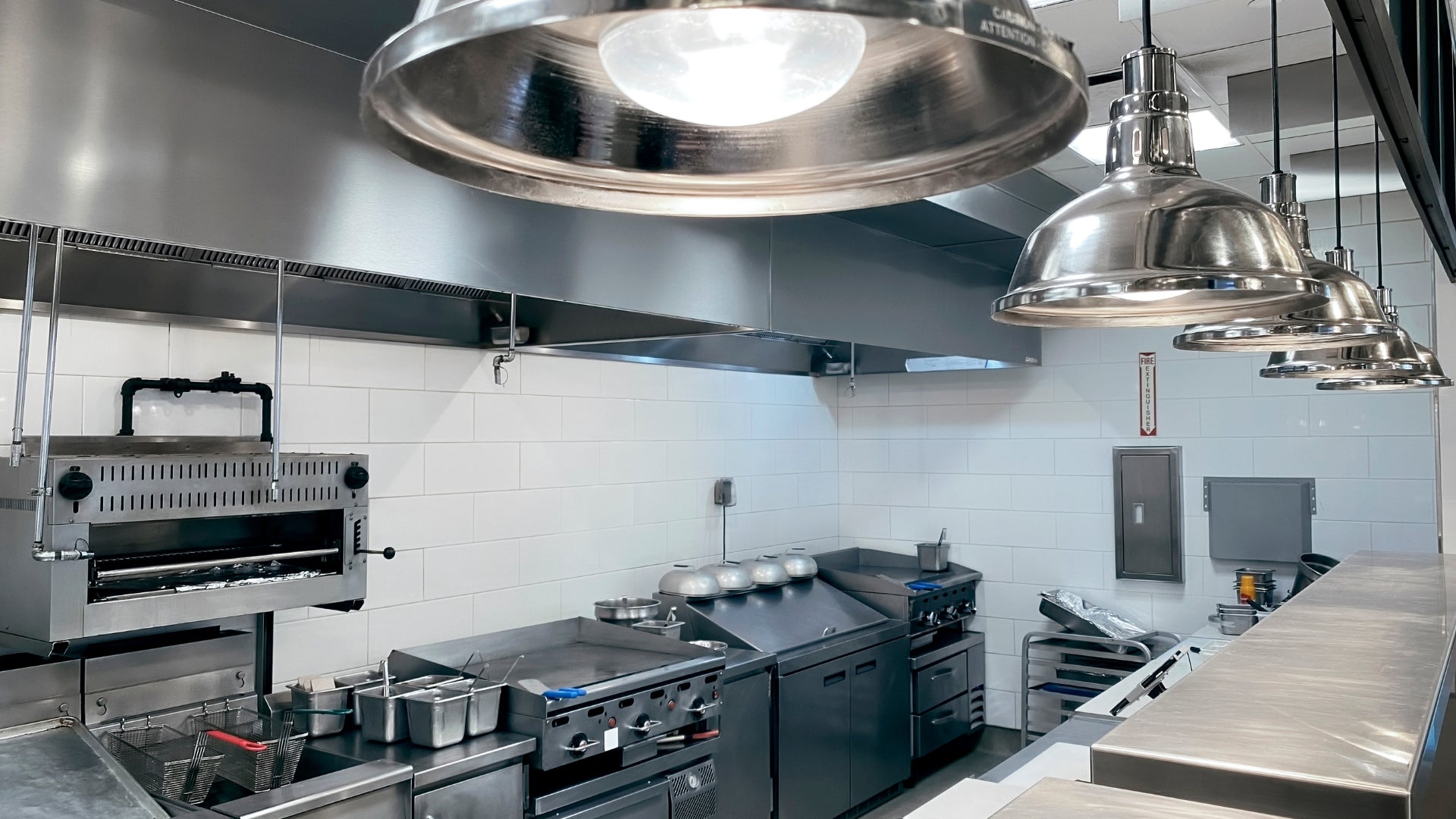 Commercial kitchen interior with stainless steel range hoods, pendant lights, counters, and cooking equipment against white tiled walls.