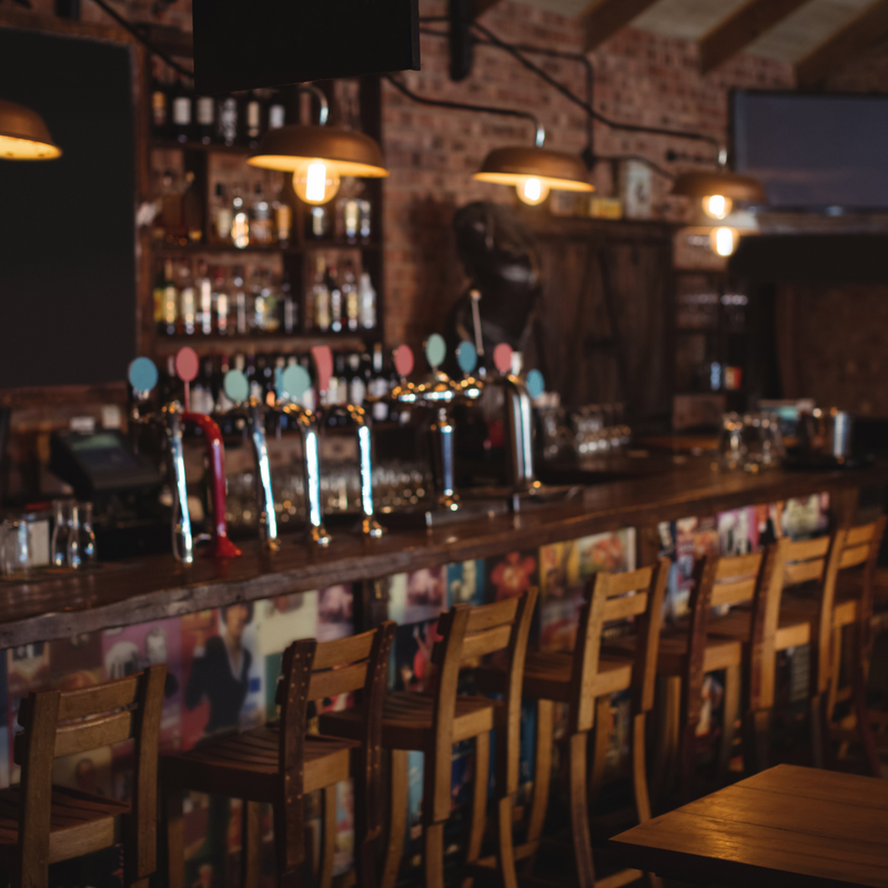 A dimly lit pub interior with a wooden bar counter, multiple beer taps with colorful handles, wooden stools, brick walls, and hanging pendant lights.