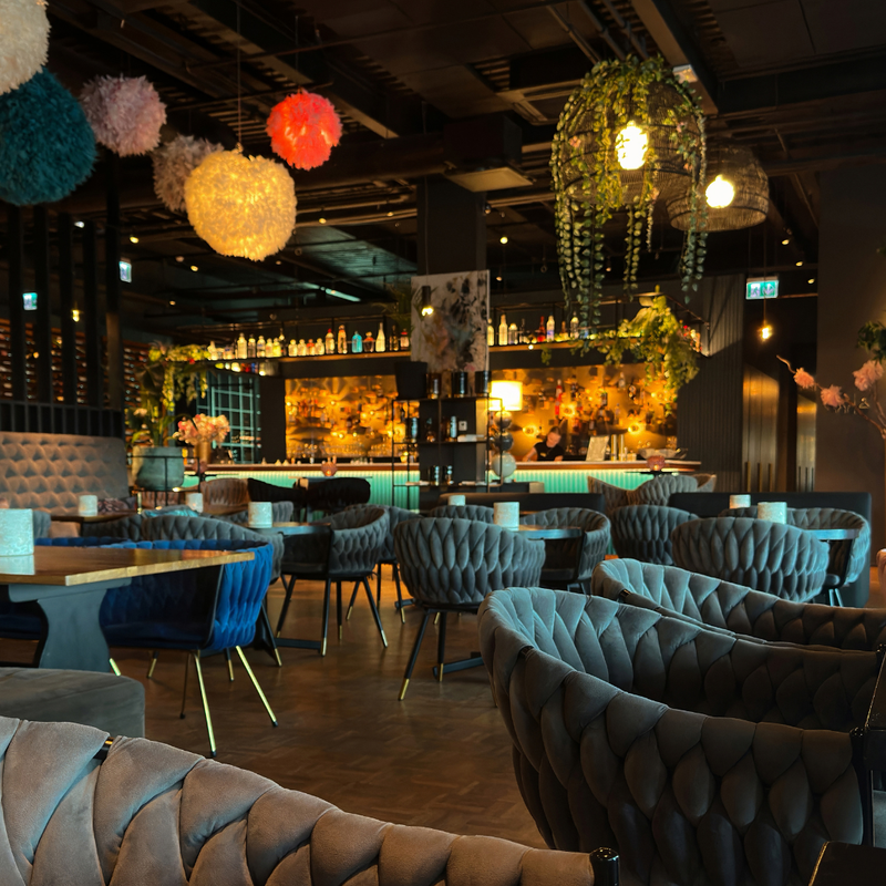 Modern restaurant interior with dark wood flooring, gray upholstered chairs, and colorful hanging lights.